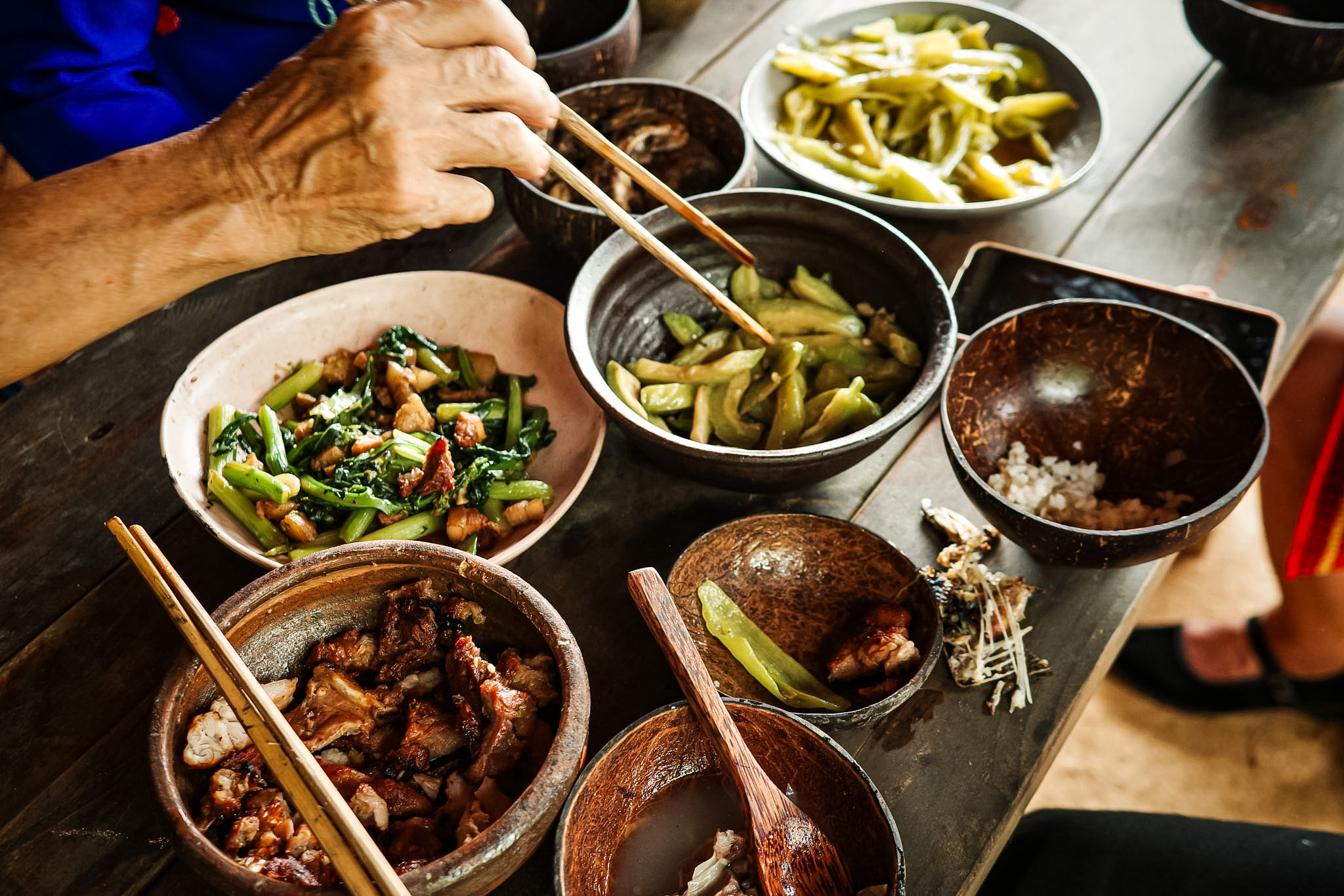 A hand with chopsticks reaching over bowls of vegetables and meat on a wooden table.