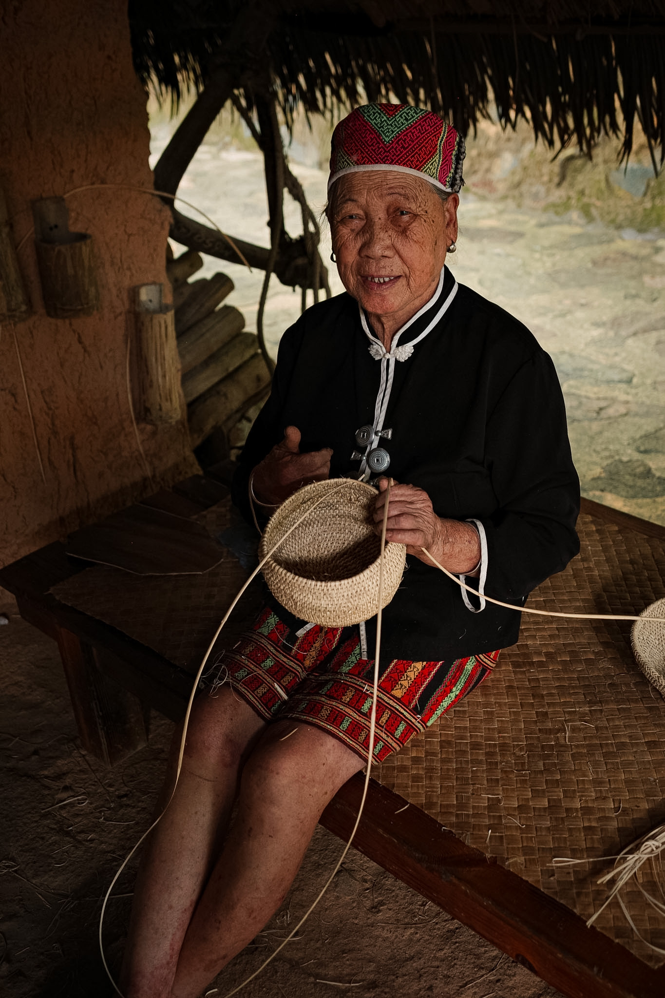 An elderly woman in traditional dress weaving a basket in front of a thatched house at Binglanggu.