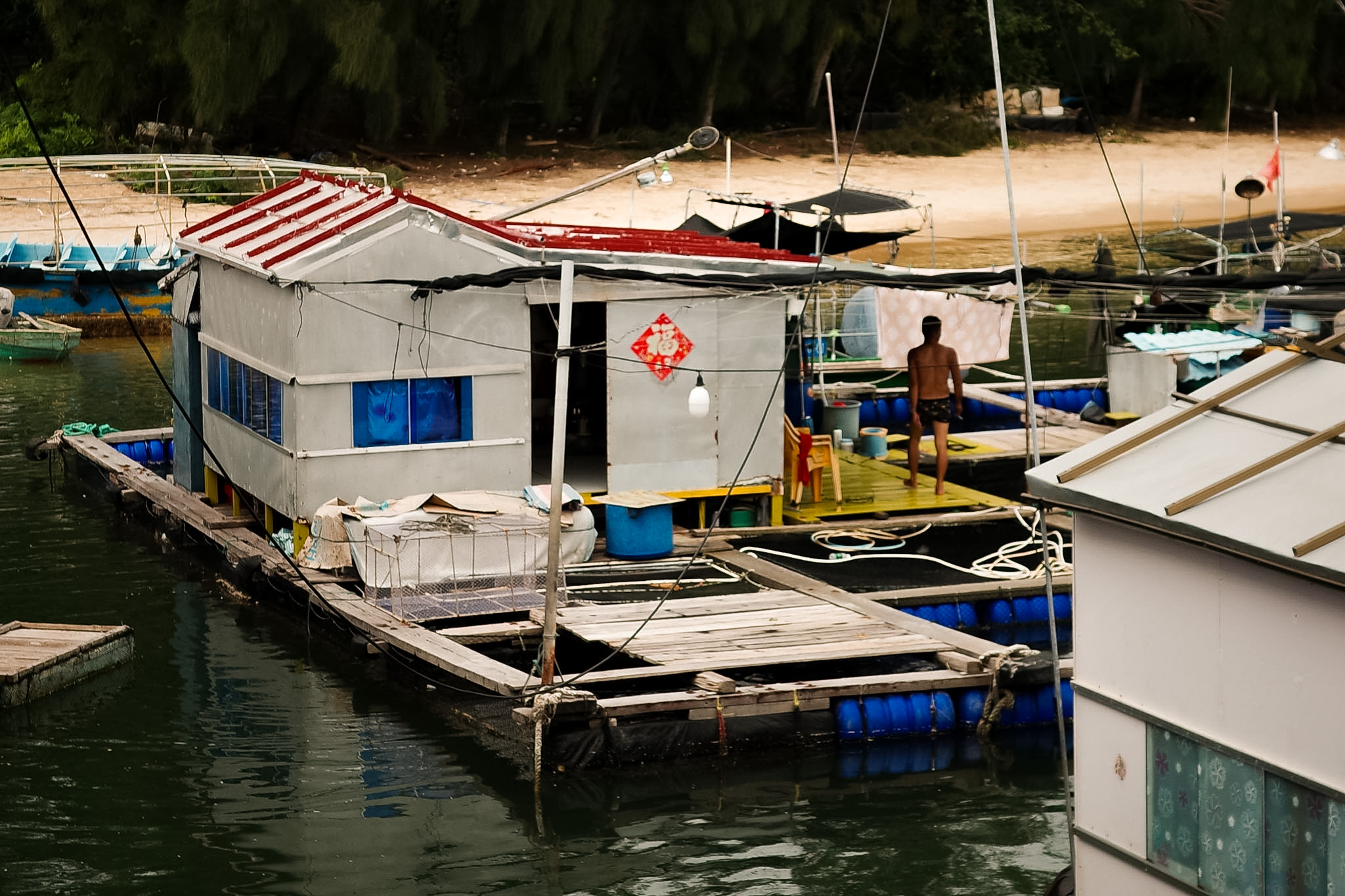 A man standing alone on the wooden deck of a floating raft house, laundry hung on a line beside him.