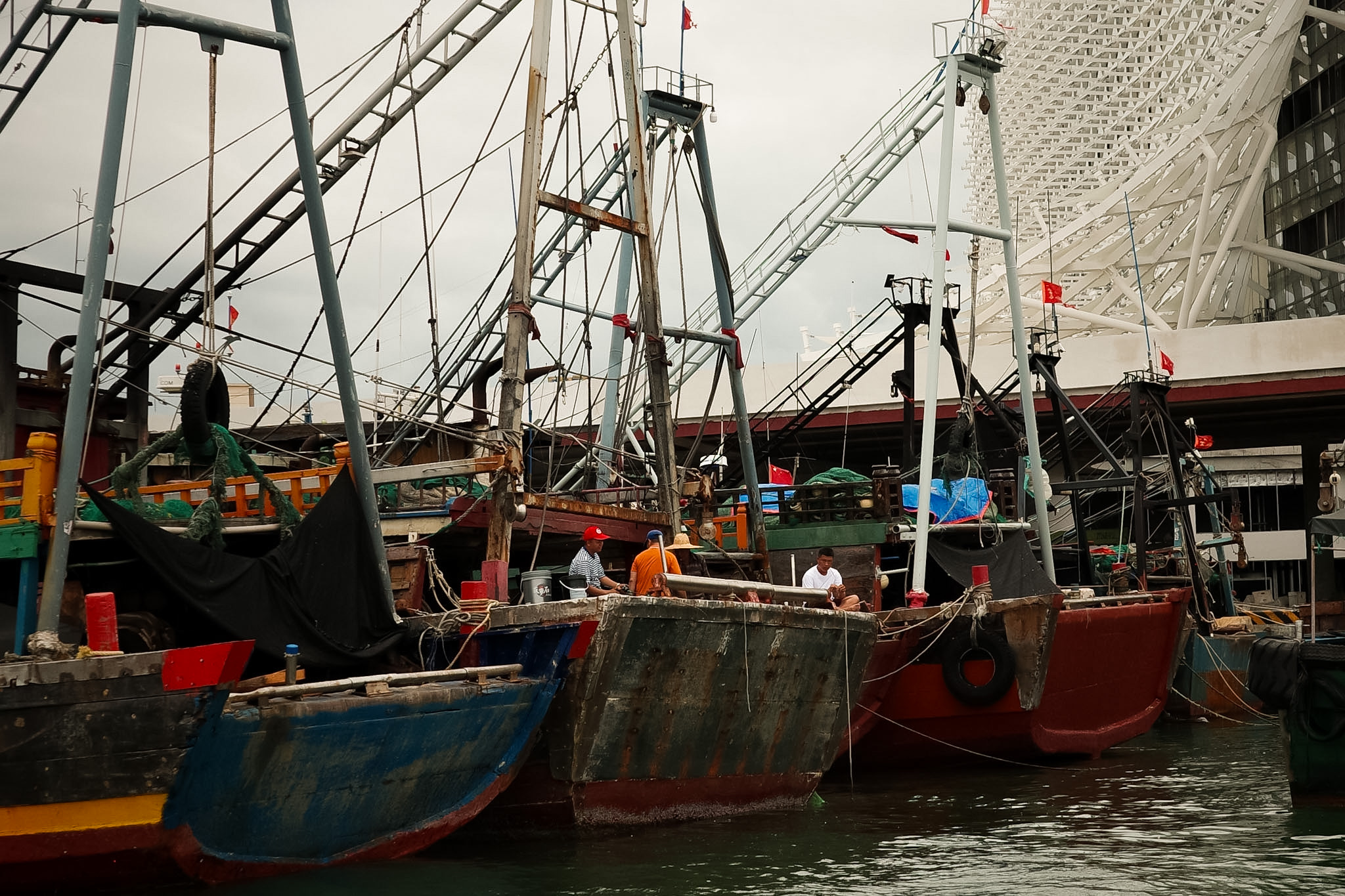 Fishing boats moored side by side, three men sitting on a bow, a white lattice building behind them.