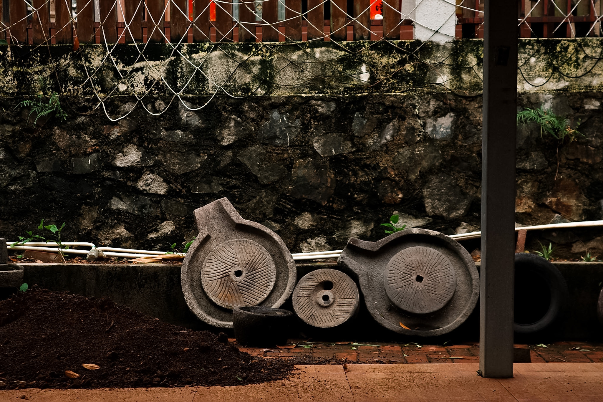 Three old stone grinding wheels leaning against a mossy stone wall beside a pile of soil.